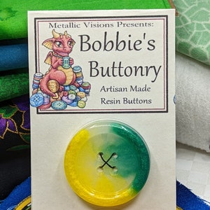 Op de afbeelding: Een gele en groene harsknop met een wit midden en een zwarte X in het midden. De knop bevindt zich op een kaart met de tekst "Metallic Visions Presents: Bobbie's Buttonry Artisan Made Resin Buttons".