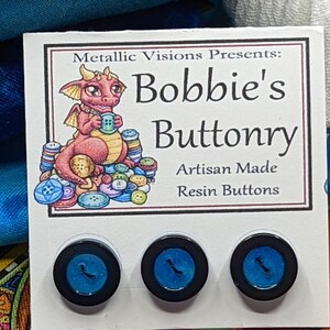 Op de afbeelding: Een kaart met een cartoon draak die een knoop vasthoudt, de tekst "Metallic Visions Presents: Bobbie's Buttonry Artisan Made Resin Buttons" en drie blauwe harsknoppen met zwarte randen.