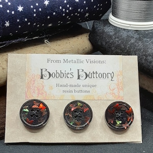 Op de afbeelding: Drie zwarte harsknoppen met een rood, oranje en geel vlinderontwerp. De knoppen bevinden zich op een kaart met de tekst "From Metallic Visions: Bobbie's Buttonry Hand-made unique resin buttons".