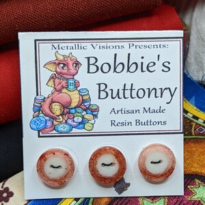 Op de afbeelding: Drie rode en witte harsknoppen met een wit centrum en een zwarte lijn in het midden. De knoppen bevinden zich op een witte kaart met de tekst "Metallic Visions Presents: Bobbie's Buttonry Artisan Made Resin Buttons".