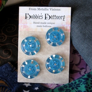 Op de afbeelding: Vier turquoise blauwe harsknoppen met een stervormig ontwerp. De knoppen bevinden zich op een witte kaart met de tekst "From Metallic Visions: Bobbie's Buttonry Hand-made unique resin buttons".