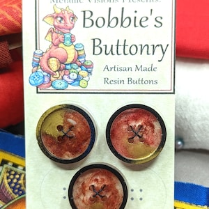 Op de afbeelding: Een kaart met een cartoon-draak die een knoop vasthoudt, de tekst "Bobbie's Buttonry Artisan Made Resin Buttons" en drie rode, gouden en zwarte knopen van hars.