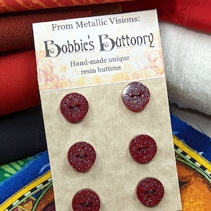 Op de afbeelding: Set van zes rode glitterharsknoppen met een zwart midden. De knoppen worden getoond op een kaart met de tekst "From Metallic Visions: Bobbie's Buttonry Hand-made unique resin buttons".