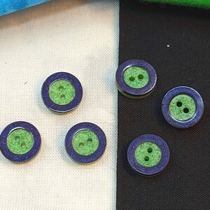 Op de afbeelding: Zes groene en paarse knopen met een ontwerp met drie gaten. De knopen hebben een groene kern met een paarse buitenring.