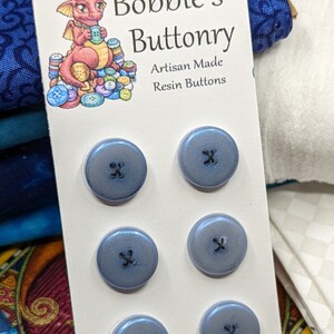 Könnte beinhalten: Eine Karte mit einem Comic-Drachen, der Knöpfe hält, dem Text "Metallic Visions Presents: Bobbie's Buttonry Artisan Made Resin Buttons" und einem Satz von vier blauen Harz-Knöpfen mit Kreuzdesign.