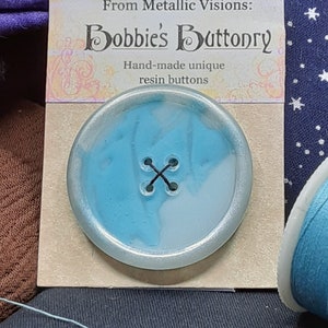 Op de afbeelding: Een lichtblauwe en witte harsknop met een zilveren rand. De knop heeft een zwarte draad die door vier gaten in het midden is genaaid. De knop bevindt zich op een kaart waarop "From Metallic Visions: Bobbie's Buttonry Hand-made unique resin buttons" staat.