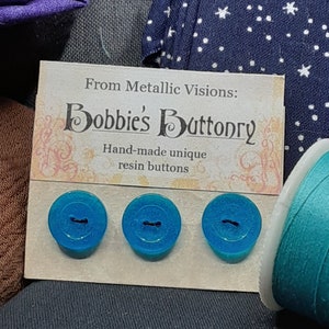 Op de afbeelding: Drie turquoise blauwe harsknoppen met een ontwerp met twee gaten. De knoppen worden gepresenteerd op een kaart waarop staat "From Metallic Visions: Bobbie's Buttonry Hand-made unique resin buttons."