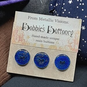 Op de afbeelding: Drie blauwe harsknoppen met een metallic afwerking. De knoppen worden gepresenteerd op een kaart met de tekst "From Metallic Visions: Bobbie's Buttonry Hand-made unique resin buttons".