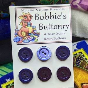 Op de afbeelding: Een kaart met een cartoon draak die een knoop vasthoudt, de tekst "Metallic Visions Presents: Bobbie's Buttonry Artisan Made Resin Buttons" en zes paarse knopen van hars.