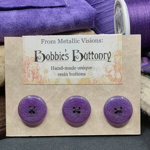Op de afbeelding: Drie paarse harsknoppen met zwarte stiksels. De knoppen worden getoond op een kaart met de tekst "From Metallic Visions: Bobbie's Buttonry Hand-made unique resin buttons."