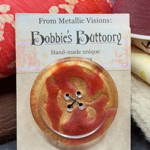 Op de afbeelding: Een gouden en rode harsknop met een zwart draadontwerp. De knop bevindt zich op een kaart met de tekst "From Metallic Visions: Bobbie's Buttonry Hand-made unique".