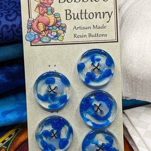 Op de afbeelding: Vijf blauwe en witte harsknoppen met een glinsterende afwerking. De knoppen hebben een wit midden met een zwarte X in het midden. De knoppen worden weergegeven op een kaart met de tekst "Bobbie's Buttonry Artisan Made Resin Buttons".