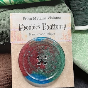 Op de afbeelding: Een handgemaakte, unieke knoop met een marmerpatroon in rood, groen en blauw. De knoop heeft een zwarte viergaats steel en is omgeven door een transparante harscoating. De knoop wordt gepresenteerd op een kaart met de tekst "From Metallic Visions: Bobbies Buttonry Hand-made unique".