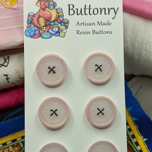 Op de afbeelding: Zes roze en witte harsknoppen met een zwarte X in het midden. De knoppen zijn gerangschikt in twee rijen van drie. De kaart luidt "Metallic Visions Presents: Bobbie's Buttonry Artisan Made Resin Buttons".