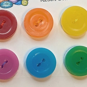 Könnte beinhalten: Sechs bunte Plastikknöpfe mit jeweils zwei Löchern. Die Knöpfe sind rot, orange, gelb, lila, blau und grün.