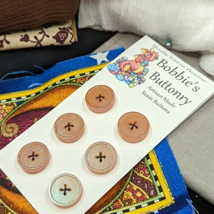 Op de afbeelding: Zes iriserende perzikkleurige harsknoppen met een kruisontwerp. De knoppen zijn gerangschikt in twee rijen van drie op een witte kaart met de tekst "Metallic Visions Presents: Bobbie's Buttonry Artisan Made Resin Buttons".