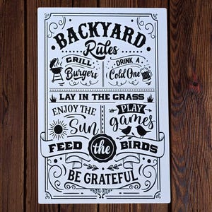 May include: A rectangular white sign with black text and illustrations. The sign reads "Backyard Rules" and includes phrases like "Grill Burgers," "Drink a Cold One," and "Feed the Birds." Decorative elements frame the text.