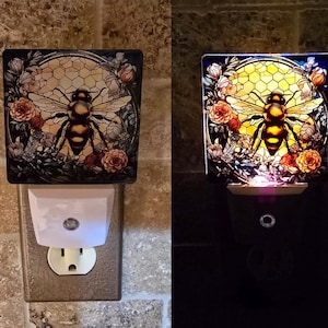 May include: A nightlight with a stained glass design of a bee in a honeycomb. The nightlight is plugged into an outlet and is lit up, casting a warm glow.