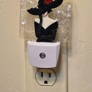 May include: A white night light with a clear glass shade featuring a black and white illustration of a woman wearing a black hat with a red flower. The night light is plugged into a standard electrical outlet.