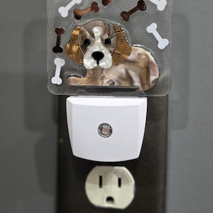 May include: A decorative night light featuring a dog design with bone accents. The night light is white and plugs into a standard electrical outlet. The dog design is brown, white, and gold.