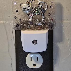 May include: A decorative night light featuring a cat figurine with floral accents. The cat is adorned with colorful jewels and sits atop a clear, glittery base with flower-shaped embellishments. The night light plugs into a standard electrical outlet.