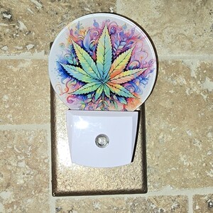 May include: A decorative night light featuring a colorful cannabis leaf design. The leaf is rendered in shades of green, yellow, orange, and blue against a swirling background. The night light has a white base and a gold-colored surround.