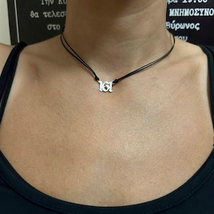 May include: A silver pendant necklace featuring the number "161" in a gothic font. The pendant is suspended from a thin, black cord. The person wearing the necklace is wearing a black top.