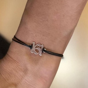 May include: A silver charm bracelet with a black cord. The charm features an abstract design. The bracelet is worn on a wrist.