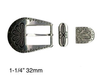 May include: A silver-toned belt buckle set with an ornate floral design. The set includes a buckle, a belt loop, and a tip. The buckle is 1-1/4" or 32mm wide. The design features detailed patterns and a vintage aesthetic.