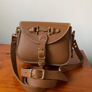 Könnte beinhalten: Eine braune Leder-Umhängetasche mit einer goldenen Schnalle und einem goldenen Metallverschluss. Die Tasche hat einen langen, verstellbaren Riemen.