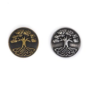 May include: Two circular metal buttons featuring a Celtic tree of life design. One is bronze with gold detailing, the other silver with a grey finish. The tree and roots are intricately detailed.