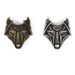 Pode incluir: Dois pingentes de cabeça de lobo de metal. Um é bronzeado com detalhes em preto, o outro é prateado com detalhes em preto. Ambos os pingentes apresentam intrincados nós celtas e desenhos geométricos.