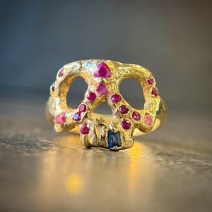 May include: A gold-colored skull ring with ruby and sapphire accents. The ring features a skull design with open eye sockets and a detailed jawline. The skull is adorned with multiple small, round, red rubies and a single blue sapphire.