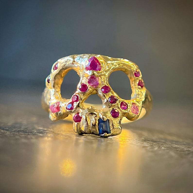 May include: A gold-colored skull ring with ruby and sapphire accents. The ring features a skull design with open eye sockets and a detailed jawline. The skull is adorned with multiple small, round, red rubies and a single blue sapphire.