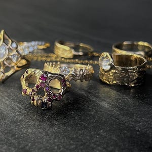 May include: A collection of gold rings with various designs, including a skull ring with red gemstones, a ring with a large clear gemstone, and rings with intricate patterns. A gold chain is also visible.