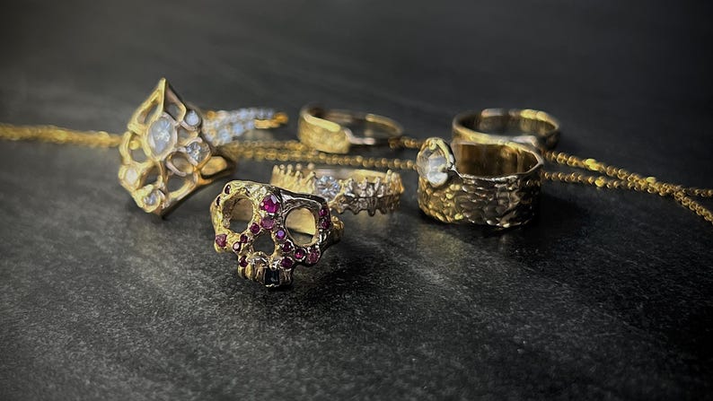 May include: A collection of gold rings with various designs, including a skull ring with red gemstones, a ring with a large clear gemstone, and rings with intricate patterns. A gold chain is also visible.