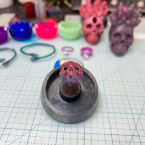 Op de afbeelding: Een close-up van een roze en teal schedelring op een metalen basis. Andere sieraden en decoratieve schedels in verschillende kleuren zijn zichtbaar op de achtergrond. De ring is het middelpunt en toont ingewikkelde details.