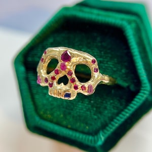 May include: A gold-colored skull ring with ruby and sapphire accents. The skull's eye sockets and nose are open, and the ring is set against a green velvet background. The ring is a unique piece of jewelry.