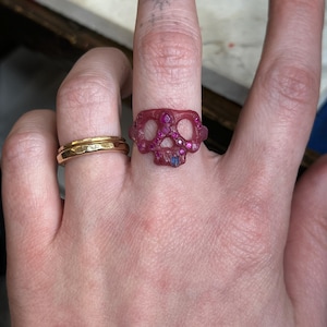 Op de afbeelding: Een roze schedelring met ingebedde roze en paarse edelstenen. De ring wordt gedragen aan een vinger naast een gouden bandring. De schedelring heeft een gedetailleerd ontwerp en is gemaakt van een doorschijnend materiaal.