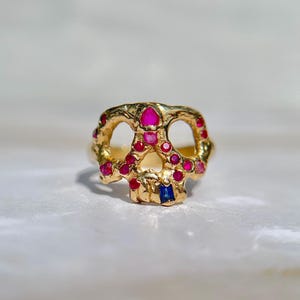 May include: A gold-colored ring in the shape of a skull, adorned with red and pink gemstones, and a single blue gemstone. The ring has a textured surface and a detailed design, showcasing a unique and artistic style.