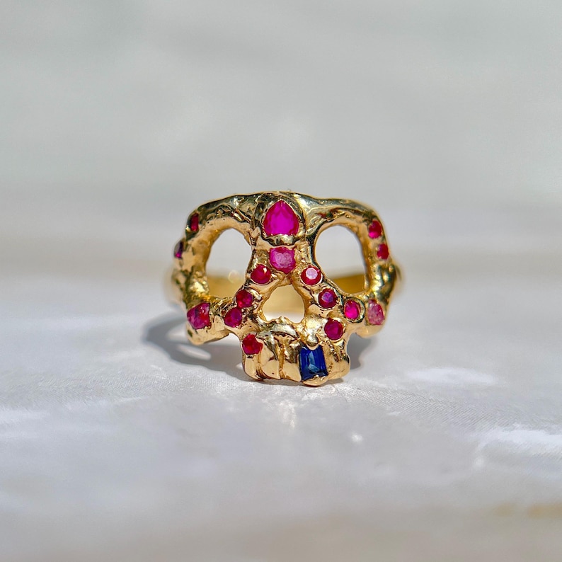 May include: A gold-colored ring in the shape of a skull, adorned with red and pink gemstones, and a single blue gemstone. The ring has a textured surface and a detailed design, showcasing a unique and artistic style.