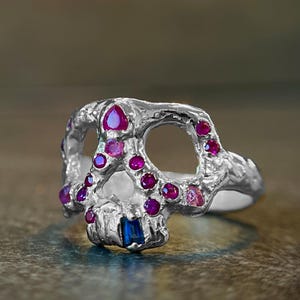 Op de afbeelding: Een zilveren schedelring met robijn- en saffieraccenten. De ring heeft een schedelontwerp met open oogkassen en is versierd met meerdere ronde en peervormige rode edelstenen en een enkele blauwe vierkante edelsteen.