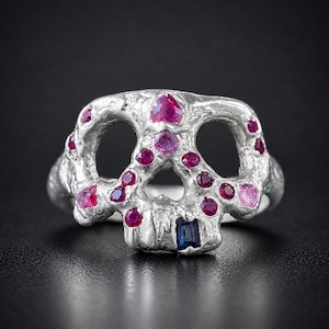 Puede incluir: Un anillo de calavera plateado con acentos de rubíes y gemas rosas. El anillo presenta un diseño de calavera con cuencas de ojos abiertas y una gema azul rectangular en la mandíbula. Las gemas están engastadas en los rasgos de la calavera.