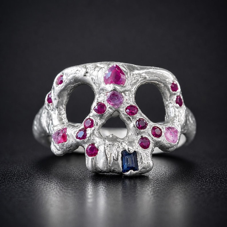 Op de afbeelding: Een zilveren schedelring met robijn en roze edelsteen accenten. De ring heeft een schedelontwerp met open oogkassen en een rechthoekige blauwe edelsteen in de kaaklijn. De edelstenen zijn gezet in de kenmerken van de schedel.