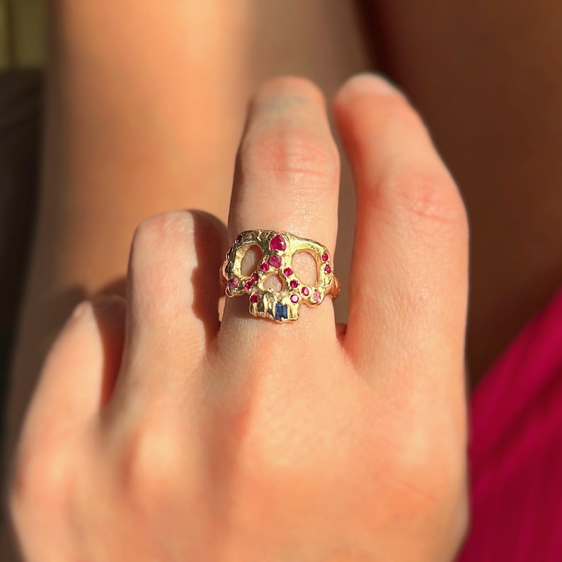 May include: A gold-colored skull ring adorned with red and blue gemstones. The ring is worn on a finger, showcasing the intricate details of the skull design and the sparkle of the gemstones.