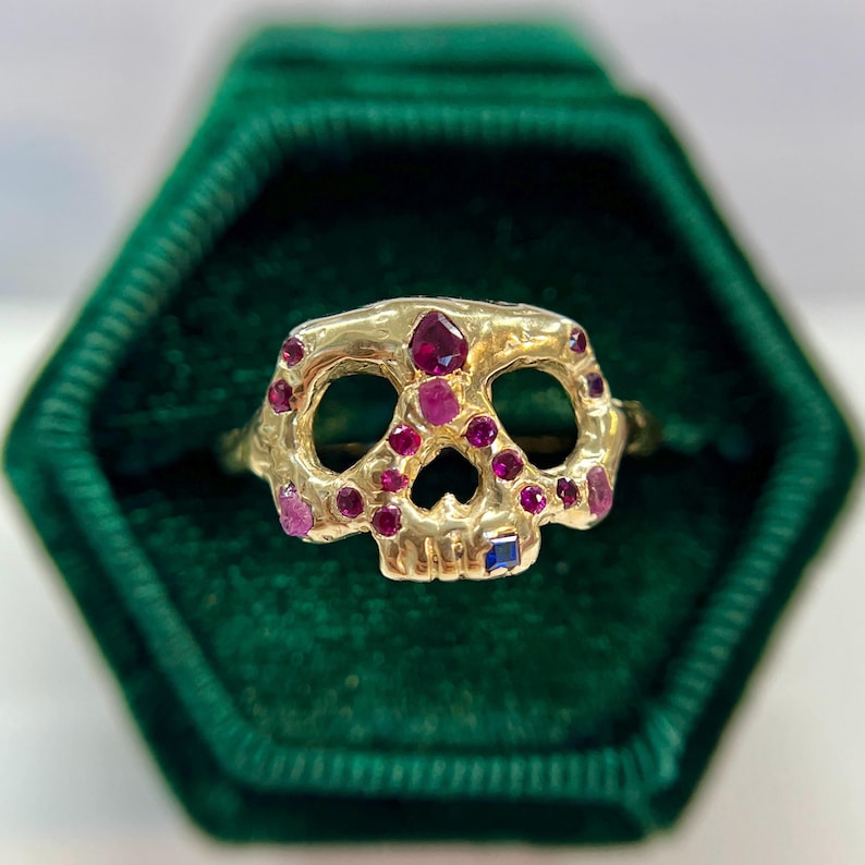 May include: A gold skull ring with ruby and sapphire accents. The skull's eye sockets and nose are open, and the ring is set with various sizes of red and pink gemstones. The ring is displayed in a green velvet box.