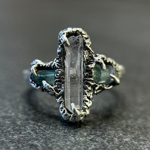May include: A silver ring featuring a central clear crystal and two blue-green gemstones. The ring has a textured, organic design, with the gemstones set on either side of the central crystal. The ring is displayed on a gray surface.