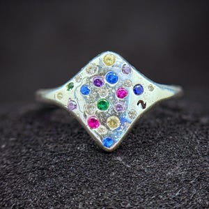 Könnte beinhalten: Ein silberner Ring mit rautenförmiger Fassung, verziert mit einer Anordnung farbiger Edelsteine. Die Edelsteine umfassen Blau, Grün, Rosa, Gelb und Lila, angeordnet in einem symmetrischen Muster. Der Ring ist vor dunklem Hintergrund dargestellt.