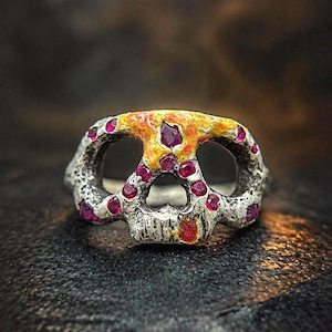 May include: A silver skull ring with ruby-colored gemstones set into the eye sockets and around the skull. The top of the skull is painted with yellow and orange enamel.