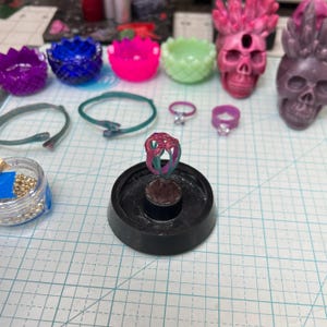 May include: A collection of jewelry and decorative items, including rings, bracelets, and skull-shaped figurines. The rings are in various colors, including pink and green. Other items include small, colorful bowls. The items are on a grid-lined surface.